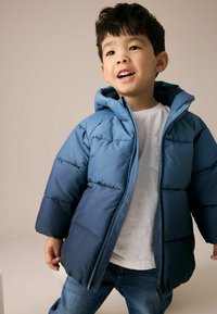 Blauwe pufferjack met capuchon, met een twee-kleurig ontwerp, geblokte textuur en ritssluiting aan de voorkant. Draagt over een witte T-shirt en denim spijkerbroek.