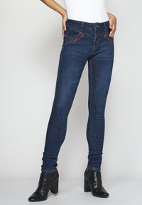 Tumman denimin kapea farkut, joissa on kukkakirjontaa taskujen lähellä ja etupuolella nappikiinnitys, yhdistettynä mustiin korkokenkiin.