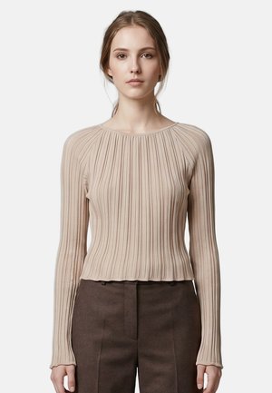 Jeune femme aux cheveux châtain clair portant un haut beige côtelé à manches longues et un pantalon marron foncé, debout avec une expression neutre.