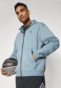 Lyseblå zipper hoodie med et sort logo på brystet, med forlommer og ribbede manchetter, parret med sorte shorts og en basketball.