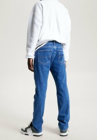 Jeans in denim blu con taglio dritto, caratterizzati da cuciture in contrasto e due tasche posteriori. Indossati con un pullover in pile bianco e sneakers.