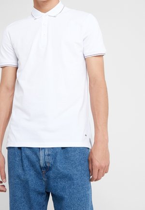 Polo blanc à trois boutons, col accentué de fines rayures foncées, associé à un jean bleu clair à coupe décontractée.