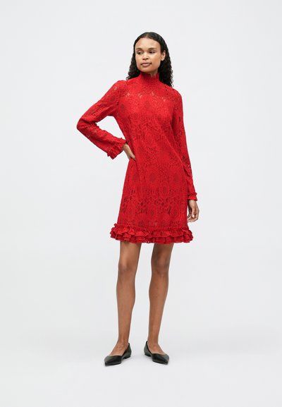 Piros csipke ruha magas nyakkal és hosszú ujjakkal, fodros szegéllyel. A dizájn virágmintákat és áttetsző részeket tartalmaz.