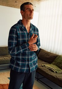 Knap-up natshirts med et grønt og blåt ternet mønster, lange ærmer og krave. Kombineret med sorte bukser i en afslappet indendørs indstilling.