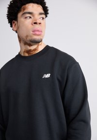 Czarna bluza z miękkiego materiału, z okrągłym dekoltem i małym białym logo New Balance na piersi, w luźnym kroju.