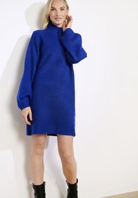 Moteris dėvi ryškiai mėlyną megztinę su pūstomis rankovėmis ir juodomis aulinėmis batais, stovinti prieš baltą foną.