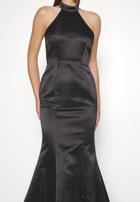 Femme portant une robe noire en satin sans manches, à col licou, avec un corsage ajusté et une jupe évasée, sur un fond blanc.