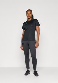 Fekete sportpóló rövid ujjakkal és fehér részletekkel, sötétszürke leggings-szel párosítva, amelyen a combon logó található és textúrás anyagú.