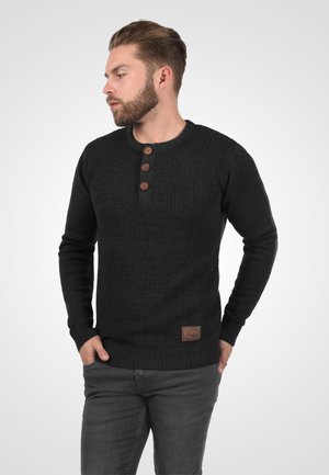 Homme barbu portant un pull en laine noir avec trois boutons marron et un pantalon gris foncé, les mains dans les poches, regardant sur le côté.