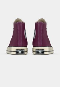 Burgundinės Converse Chuck Taylor All Star aukštakulnės sportinės avalynės pora rodyta iš nugaros ant balto fono.