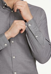 Camicia grigia con bottoni a pressione e tessuto testurizzato. Presenta bottoni bianchi e maniche arrotolabili. Il design include un colletto e una silhouette aderente.