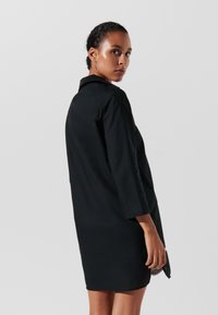 Abito camicia nero con colletto, maniche lunghe e taglio dritto. Presenta una texture liscia e arriva sopra il ginocchio.
