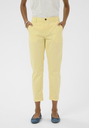 Naine kannab lühikesi kahvatukollaseid pükse, valget topi, siniseid baleriinasussisid ja hõbedast käevõru, käed taskutes, seisab valge tausta ees.