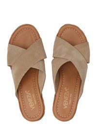 Beige suède slippers met gekruiste banden, leren binnenzool en contrasterende stiksels. De zool is plat met een afgeronde teen ontwerp.
