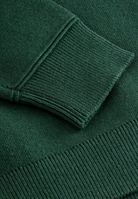 Dunkelgrüner, gestrickter Pulloverstoff mit gerippten Bündchen und Saum, zeichnet sich durch eine weiche Textur und ein einheitliches Muster mit minimal sichtbaren Nähten aus.