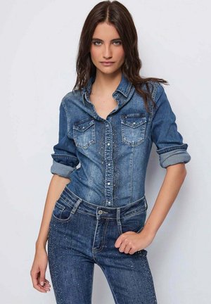 Giovane donna con lunghi capelli scuri, indossa una camicia di jeans aderente e jeans con le maniche arrotolate e un dettaglio sottile a macchie.