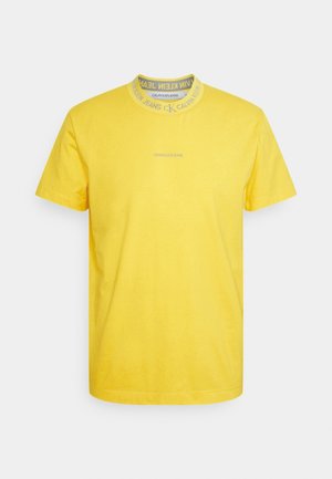Gelbes T-Shirt mit kurzen Ärmeln und Calvin Klein Jeans Logo auf der Brust sowie wiederholtem Branding rund um den Rundhalsausschnitt.