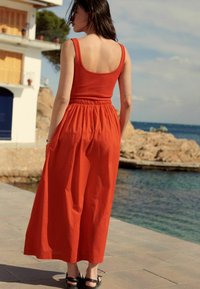 Un vestido rojo sin mangas con un top ajustado y cintura elástica, que fluye hacia una falda larga. Combinado con sandalias negras, en un entorno al aire libre.