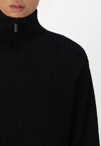 Gros plan sur une personne portant un pull en tricot côtelé noir avec un col zippé haut.