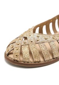 Sandalia de purpurina dorada con un diseño de tiras y tachuelas doradas. Punta abierta, plantilla acolchada y suela marrón. Material suave y texturizado.