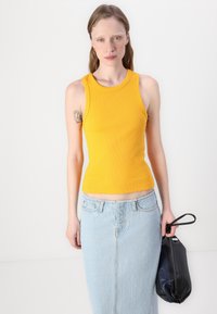 Débardeur jaune côtelé avec une coupe ajustée, associé à une jupe en denim bleu clair. Le mannequin tient un sac à main noir. Texture lisse sur l'ensemble.