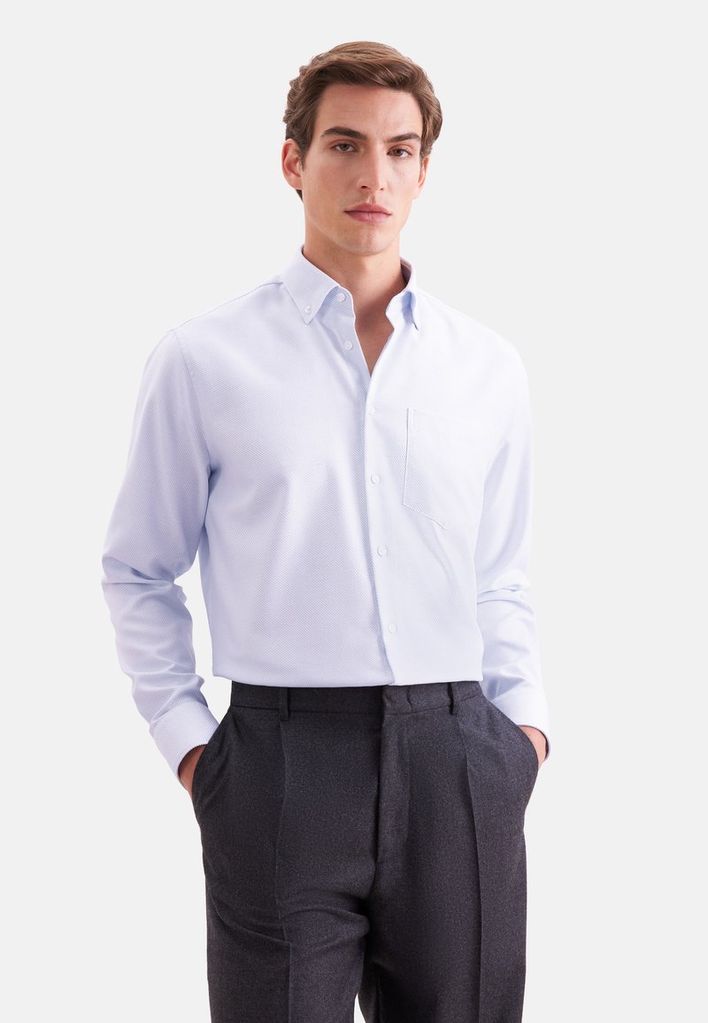 Jeune homme portant une chemise bleu clair boutonnée rentrée dans un pantalon gris foncé, debout les mains dans les poches devant un fond uni.