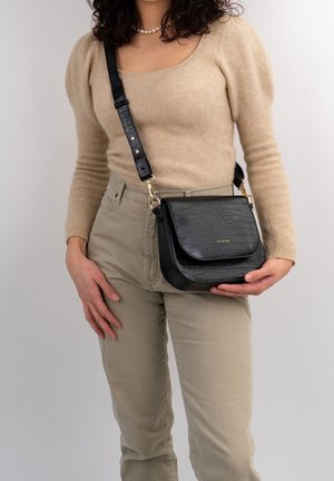 Nainen, joka pukeutunut beigeen neulepaitaan ja housuihin, pitää kädessään mustaa krokotiilikuvioista olkalaukkua, jossa on kultaiset metalliosat ja säädettävä hihna.