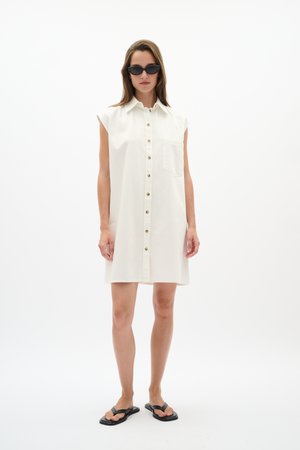 Kobieta ubrana w białą sukienkę bez rękawów zapinaną na guziki z kieszenią z przodu, czarne okulary przeciwsłoneczne w stylu cat-eye i czarne płaskie sandały, stojąca na białym tle.