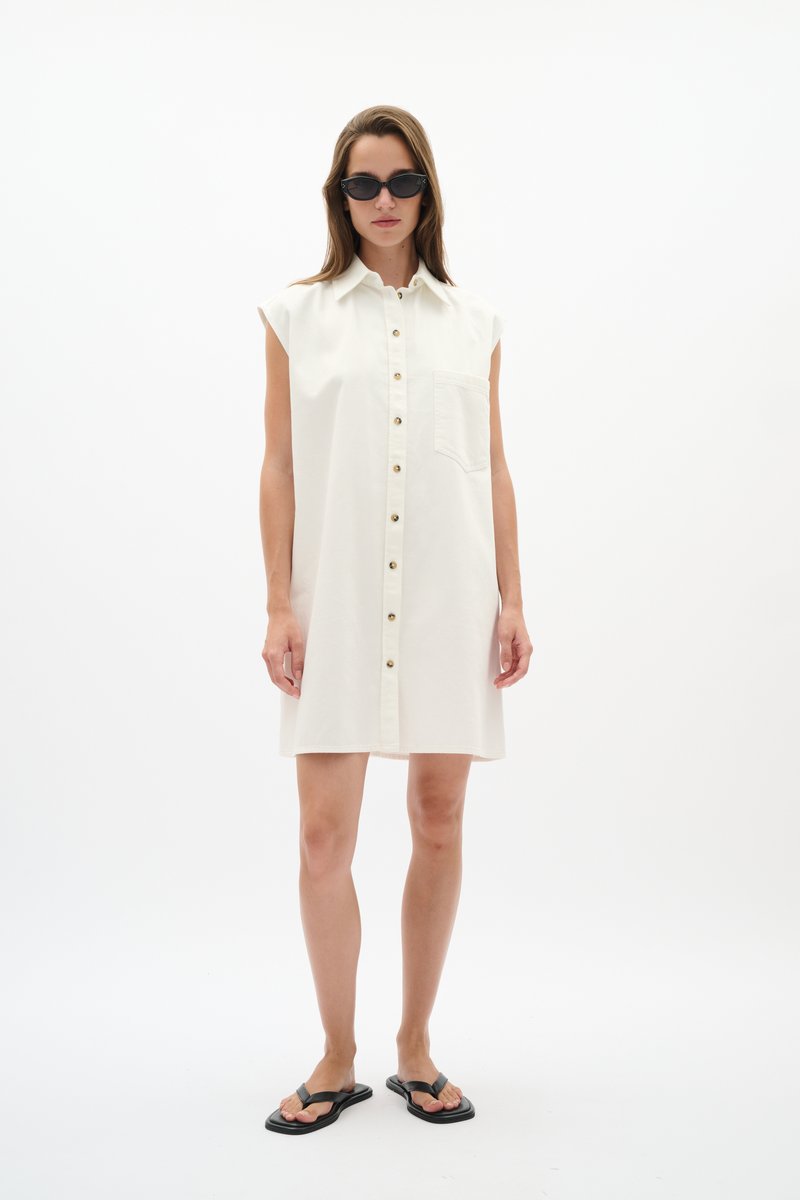 Femme portant une robe blanche sans manches boutonnée devant avec poche avant, lunettes de soleil noires style œil de chat et sandales plates noires, debout sur fond blanc.
