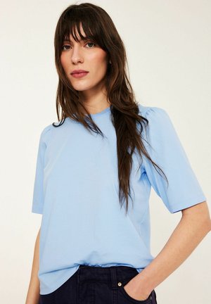 Młoda kobieta z długimi brązowymi włosami, ubrana w jasnoniebieską bluzkę z krótkim rękawem i ciemne spodnie, stoi na tle jednolitego jasnego tła.