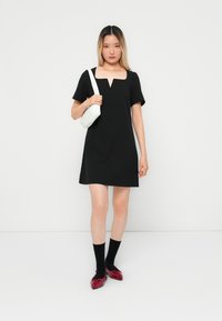 Black short-sleeve dress with a square neckline and inverted triangle cutout detail; paired with red shoes and black socks, holding a white bag.