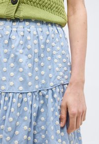 Δεξί χέρι και χέρι δίπλα σε μια ανοιχτό μπλε φούστα με λευκές πουά και ένα πράσινο πλεκτό πουλόβερ με ένα μαύρο κουμπί.