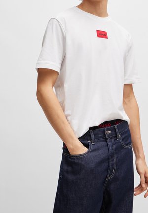 Person trägt weißes kurzärmeliges T-Shirt mit rotem "HUGO"-Logo und dunkelblaue Jeans, Hand in der Tasche, vor einfarbig hellem Hintergrund.