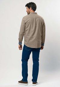 Mann, der ein braunes Langarmhemd über blauem, umgeschlagenem Hemd, blauen Jeans und braunen Turnschuhen trägt, steht mit dem Rücken zur Kamera vor einem weißen Hintergrund.