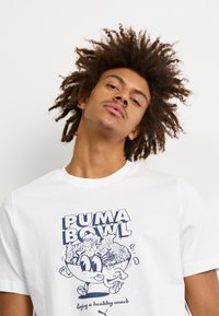 Balta medvilninė trumpa rankovė T-shirt su grafiniu atvaizdu, kuriame pavaizduota dubuo su maistu ir užrašas "PUMA BOWL" mėlyna spalva, apvali iškirptė.