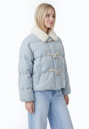 Ung kvinde iført en lyseblå dunjakke med knaplukning og hvidt imiteret pelskrave, kombineret med lyseblå jeans.