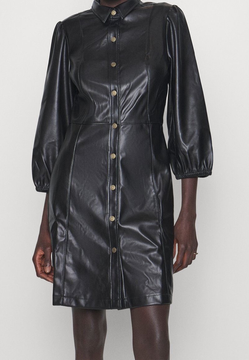 Robe noire en simili cuir longueur genou avec manches longues bouffantes, fermeture boutonnée à l'avant et col pointu portée par une personne debout.