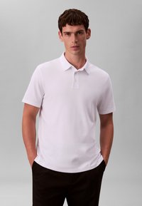 Tricou polo alb cu mâneci scurte, guler clasic și fenta cu trei nasturi, realizat dintr-un material neted, asortat cu pantaloni negri.