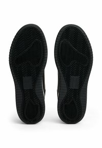 Suole di scarpe in gomma nera con un motivo a zigzag, caratterizzate da un design piatto e bordi lisci. Presentano un branding minimale nella zona del tallone.