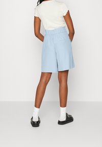 Štíhlá osoba v bílém tričku s krátkým rukávem, světlomodrých šortkách s vysokým pasem, bílé ponožky a černé slip-on boty s hroty.