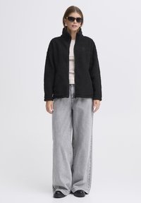 Must musternakk, millel on kõrge krae, on kombineeritud heleda beeži pluusi, hallide laia jalgadega teksade ja mustade kingadega. Mudel kannab prille.