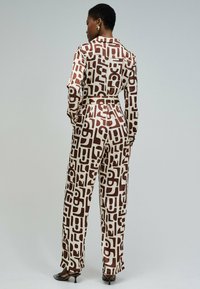 Tuta marrone e crema con motivi, maniche lunghe, collo, vita con cintura e gambe larghe. Tessuto liscio con forme astratte e accenti.