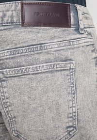Jeans de denim gris claro con un acabado desgastado. Presenta un parche de cuero marrón etiquetado como "RIVER ISLAND" y bolsillos traseros estándar.