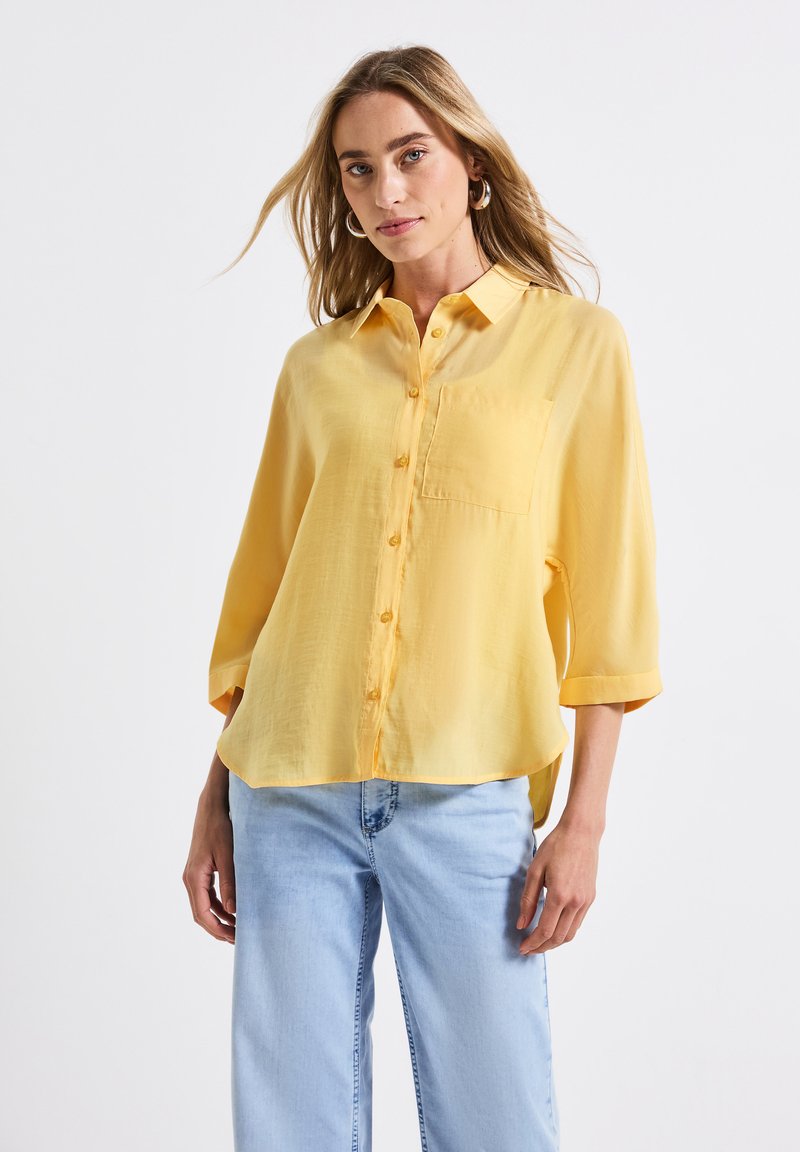 Camicia gialla con bottoni, realizzata in tessuto leggero, dotata di tasca sul petto, maniche corte e orlo curvo. Abbinata a jeans azzurri chiari.