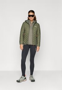 Warmes olivgrünes Pufferjackett mit gestepptem Design, grauen Lagen darunter, schwarzen Leggings und grauen Sportschuhen mit pinken Akzenten.