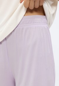 Κοντινό πλάνο χεριού που σηκώνει λευκό πουκάμισο για να αποκαλύψει ελαστική μέση ανοιχτό μωβ παντελονιού με διακριτικό ανάγλυφο λογότυπο στη πλευρά.