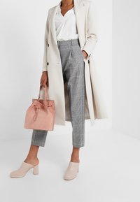 Beige lange jas, witte blouse, grijs geruite broek, roze suède handtas met trekkoord en lichtroze blokhak muilen.