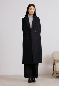 Cappotto nero doppiopetto con un design strutturato, silhouette lunga, texture liscia e grandi bottoni, indossato sopra un dolcevita grigio.