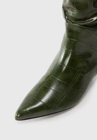 Stivale alla caviglia verde scuro a punta con texture di coccodrillo e finitura lucida su sfondo bianco.