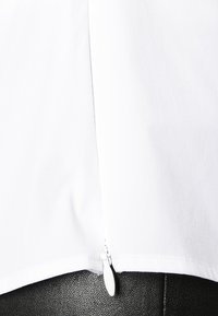 Camicia bianca a maniche corte con una texture liscia. Presenta un dettaglio con zip all'orlo con un cursore di colore argento, abbinata a pantaloni neri.